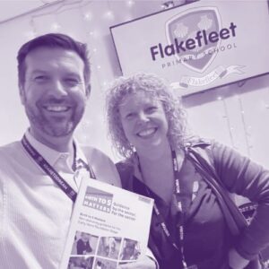 Ruth and Dave McParlin stood in front of the Flakefleet Primary School logo holding a copy of 'Birth to 5 Matters'